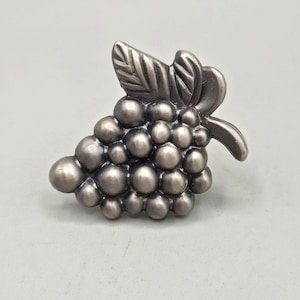 May include: A metallic silver-toned cabinet knob shaped like a cluster of grapes with a single leaf at the top. The surface has a smooth, brushed finish with rounded, spherical details representing individual grapes in the bunch.