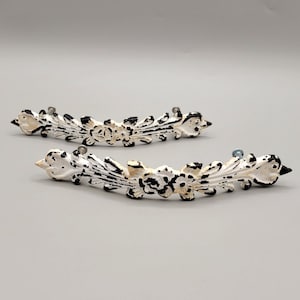 May include: Two white and black ornate metal drawer pulls with a floral design.