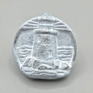 May include: A white and gray lighthouse relief sculpture. The lighthouse is centered within a circular frame, with waves and rocks at the base. The sculpture has a textured, weathered appearance.