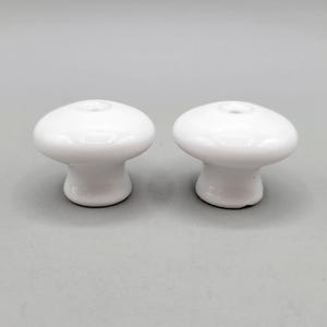 Peut inclure: Deux boutons de meuble blancs en céramique en forme de champignon. Chaque bouton a un dessus arrondi avec un petit trou et une base courte et cylindrique. Les boutons sont brillants et réfléchissent la lumière, suggérant une finition lisse. Ces boutons conviennent aux meubles ou aux projets décoratifs.