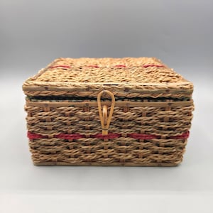 May include: A rectangular woven basket with a hinged lid, featuring natural tan fibres and horizontal bands of red cord. The basket includes a central vertical loop fastener made of the same material, securing the lid to the main body.