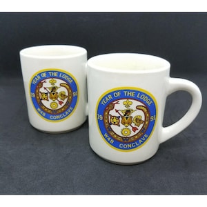 May include: Two white ceramic mugs with a blue and yellow design. The design features a lodge emblem with the text "Year of the Lodge 1991 WAB Conclave".