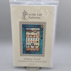 May include: A Cactus Cat Patterns quilt pattern titled "Happy Trails." The pattern features a Southwest-themed design with colorful buildings, cacti, and geometric patterns. The finished quilt size is 50 x 70 inches.