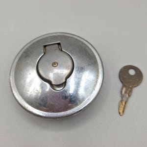 May include: A silver-colored, circular fuel tank cap with a hinged, smaller cap and a key. The key is small, with a circular hole at the top and a ridged edge. The cap has a slightly worn appearance.