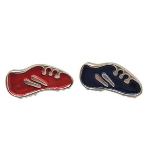 May include: Two shoe-shaped pins. One is red with silver accents, and the other is navy blue with silver accents. The pins have a glossy finish and are set against a white background.
