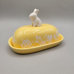 May include: A yellow ceramic butter dish with a white floral design and a white bunny figurine on top. The dish has a lid and a base, perfect for serving butter. The bunny is sitting upright.