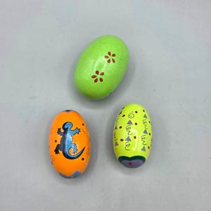 May include: Three painted wooden egg-shaped toys. One is bright green with red flower accents. Another is orange with a blue lizard design. The third is yellow with red triangles and green swirls.