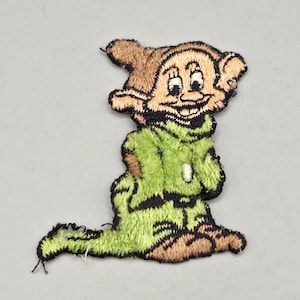 May include: Embroidered patch featuring a cartoon character in a green outfit and brown hat. The patch has a smiling face with black outlines and details. The character is sitting down with its legs extended.