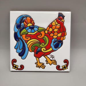 May include: A square, white ceramic tile featuring a colorful rooster design. The rooster is depicted in vibrant shades of red, orange, yellow, green, and blue, with decorative floral accents. The tile measures approximately 6 inches by 6 inches.