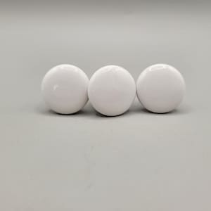 May include: Three glossy, white, spherical cabinet knobs are arranged in a row. The knobs have a smooth, reflective surface and are evenly spaced against a neutral gray background. These knobs are suitable for furniture or home decor projects.