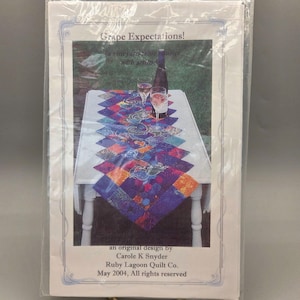 May include: A packaged quilt pattern titled "Grape Expectations!" featuring a vineyard-themed table runner. The runner has a colorful, geometric design with purple, blue, and orange hues. The image includes a bottle of wine and two glasses.