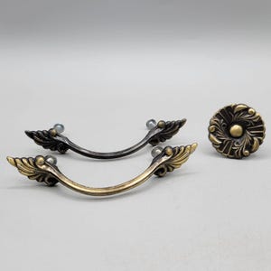 May include: Decorative cabinet hardware set featuring two ornate pull handles and a round knob. The handles are in antique brass and black finishes, with a leaf design. The knob has a floral pattern and a matching antique brass finish.