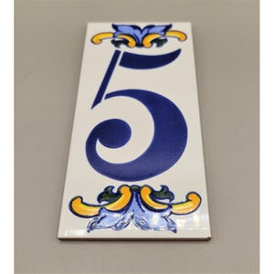 May include: Rectangular white ceramic tile with the number 5 in dark blue. The tile is decorated with blue, yellow, and green floral accents at the top and bottom. The tile is likely for a house number.