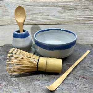 May include: A matcha tea set featuring a white ceramic bowl with a blue rim, a bamboo whisk, a bamboo scoop, and a small ceramic holder with a wooden spoon. The set is arranged on a gray stone surface.