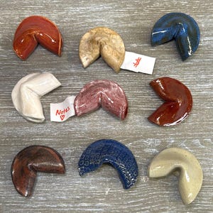 May include: Ten ceramic fortune cookies in various colors and patterns, including red, brown, white, beige, blue, and pink. The cookies are arranged in a semi-circle on a wooden surface.