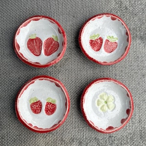May include: Four small ceramic dishes with red rims. Two dishes feature a pair of red strawberries with green leaves. One dish has a single green flower. The remaining dish is blank.