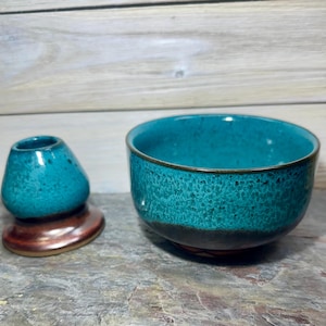 May include: A teal ceramic tea bowl and a matching whisk holder. The bowl has a textured surface with a dark brown rim and base. The whisk holder is teal with a brown base. The items are displayed on a gray stone surface.