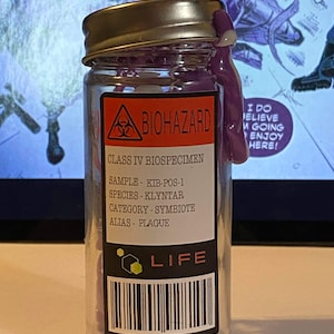 May include: A clear glass jar with a purple liquid inside. The jar has a black and white label with the words "BIOHAZARD", "CLASS IV BIOSPECIMEN", "SAMPLE - KIB-POS-1", "SPECIES - KLYNTAR", "CATEGORY - SYMBIOTE", "ALIAS - PLAGUE", and "LIFE".