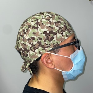 Op de afbeelding: Een persoon draagt ​​een chirurgische muts met camouflagepatroon, een blauw chirurgisch masker en een zwarte bril.