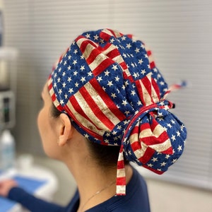 May include: A blue and red surgical scrub hat with a stars and stripes pattern. The hat is tied at the back with a long tie.