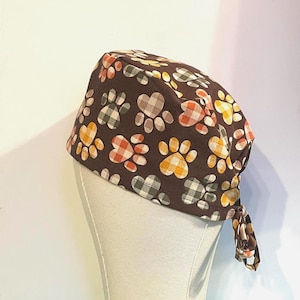 Puede incluir: Un gorro quirúrgico marrón con un patrón repetido de huellas de patas en amarillo, naranja y verde. Las huellas tienen un diseño de cuadros. El gorro está atado en la parte posterior.