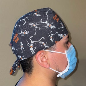 May include: A black scrub hat with a pattern of skeletons dancing and the text "Grateful Dead". The hat has a tie on the back. A person is wearing the hat and a light blue surgical mask.