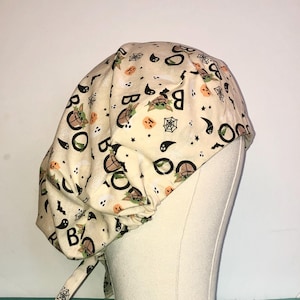 May include: A beige scrub hat with a Halloween-themed print featuring a cartoon character, pumpkins, bats, and the word "Boo".