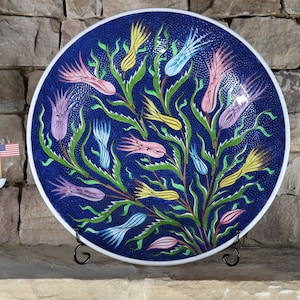 May include: A large, round ceramic plate with a blue background and a floral design. The flowers are painted in shades of pink, yellow, and blue. The plate is on a black stand.