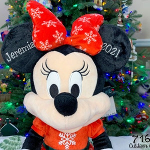 May include: A red and white plush Minnie Mouse Christmas ornament with a red bow and snowflake pattern. The ornament is personalized with the name "Jeremia" and the year "2021".
