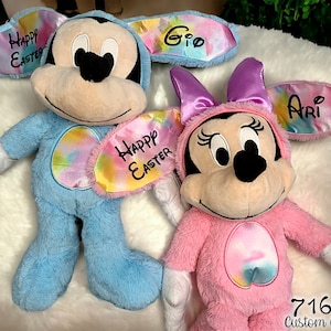 May include: Two plush Easter bunny toys, one blue and one pink, with tie-dye patterns and personalized names. The blue bunny has a yellow eye mask with the text "Happy Easter Gio" and the pink bunny has a purple bow and a yellow eye mask with the text "Happy Easter Ari".