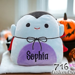 May include: A plush blue and purple vampire toy with a red cape and black bow tie. The toy has a stitched-on face with black eyes and a small mouth with fangs. The toy is personalized with the name "Sophia".