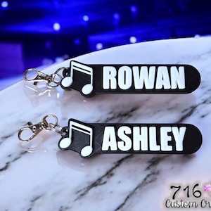 May include: Two black keychains with white text that reads "ROWAN" and "ASHLEY". Each keychain has a silver clasp and a musical note design. The background is a blurred blue and white.