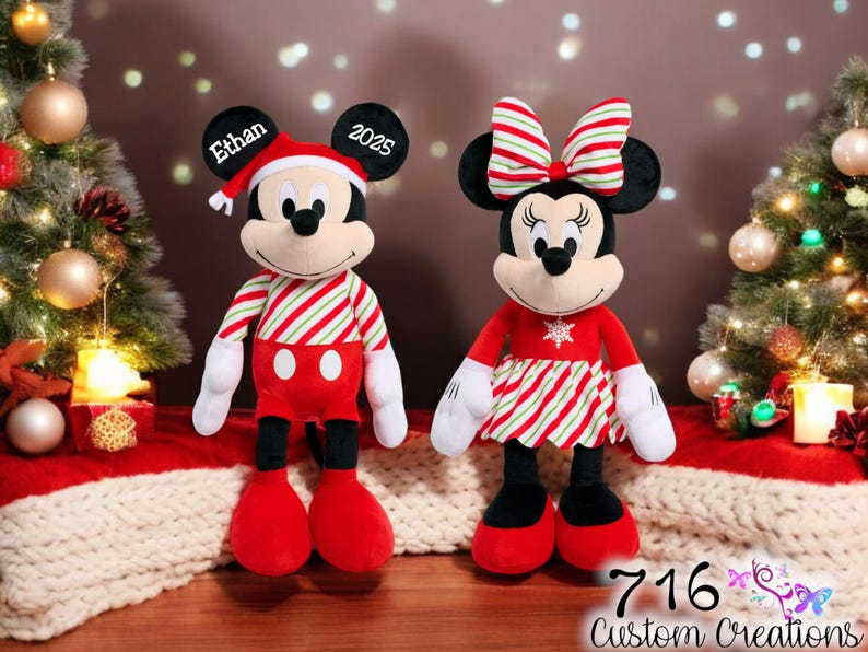 May include: Personalized Christmas Mickey and Minnie Mouse plush toys. Mickey is wearing a red and white striped shirt and red pants. Minnie is wearing a red dress with a white snowflake and red and white striped trim. Mickey's ear says "Ethan 2025".