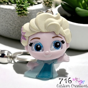May include: A blue and white figurine keychain of Elsa from the movie Frozen. The figurine has sparkly blue eyes and a light pink face. The keychain is attached to a silver ring.