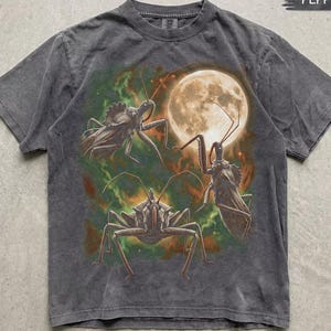Op de afbeelding: Een antracietgrijs t-shirt met korte mouwen en een grafische print van drie grote insecten tegen een achtergrond van een gloeiende volle maan en wervelende groene en oranje nevelwolken. Het shirt heeft een ronde hals en een vervaagde textuur.