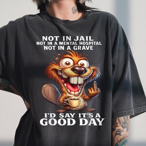 以下が含まれることがあります： コーヒーカップを持ち、中指を立てた漫画のビーバーが描かれた濃い灰色のTシャツ。「NOT IN JAIL, NOT IN A MENTAL HOSPITAL, NOT IN A GRAVE, I'D SAY IT'S A GOOD DAY」と書かれたテキスト。