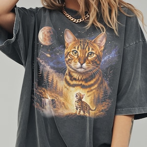 May include: Dark gray t-shirt featuring a large, detailed illustration of a Bengal cat with a smaller kitten. The design includes a forest, waterfall, moon, and starry sky. The shirt has short sleeves and a relaxed fit.