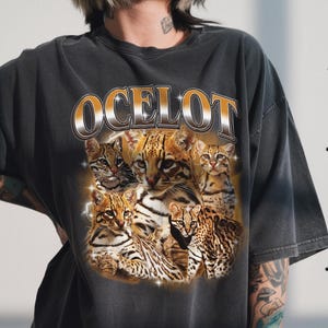 Może przedstawiać: Ciemnoszara koszulka z grafiką przedstawiającą kilka ocelotów i napisem "OCELOT" w kolorze złotym i białym. Oceloty mają brązowe i czarne cętkowane futro. Koszulka ma wygląd vintage.
