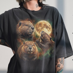 May include: Dark gray t-shirt featuring a graphic of three beavers with a full moon in the background. The beavers are depicted in shades of brown and tan, with detailed fur and expressive faces. The moon is a warm yellow color, and the background has green and orange hues.