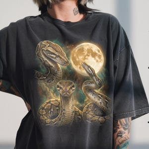 May include: Dark grey t-shirt with a graphic of three snakes and a full moon. The snakes are brown and tan with detailed scales. The moon is a warm yellow colour, and the background has a cloudy, ethereal effect.