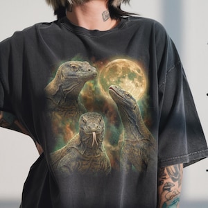 May include: Dark gray t-shirt featuring a graphic of three Komodo dragons with a full moon in the background. The dragons are detailed with realistic scales and textures, set against a colorful, cloudy sky.