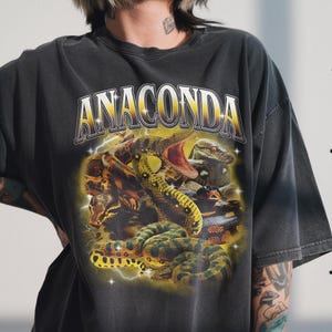 Peut inclure: T-shirt gris foncé avec le mot "ANACONDA" en lettres dorées. En dessous, un motif de plusieurs serpents verts, jaunes et bruns, sur fond d'éclair jaune.