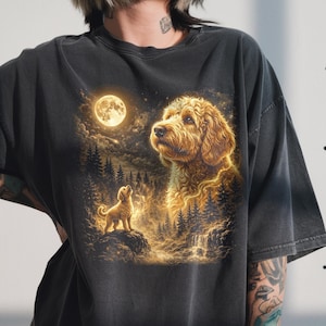 May include: Dark grey t-shirt with a golden retriever illustration under a full moon. The design includes a smaller dog on a rock, trees, and a waterfall, creating a dreamy, nature-inspired scene.