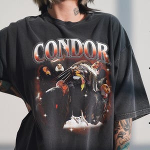 Puede incluir: Camiseta gris oscuro con la palabra "CONDOR" en fuente retro roja y blanca. La camiseta presenta un gráfico de varios cóndores con un fondo estrellado. La camiseta tiene un aspecto vintage y desgastado.