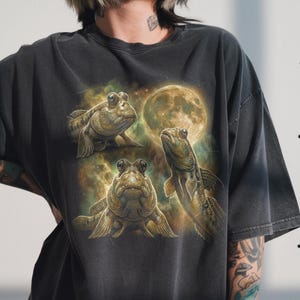 May include: Dark gray t-shirt featuring a detailed illustration of three mudskipper fish with a full moon in the background. The artwork has a vintage, distressed look, with a blend of earthy tones and a celestial theme.