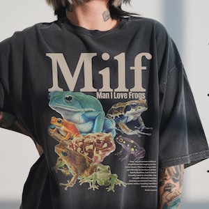 May include: Dark grey t-shirt featuring a graphic of various colourful frogs and the text "Milf Man I Love Frogs". The shirt has a vintage, distressed appearance.
