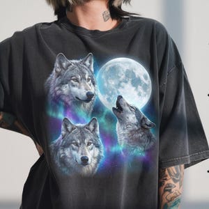 May include: Dark gray t-shirt featuring a graphic of three wolves and a full moon. The wolves are depicted in shades of gray and white, with a howling wolf in the center. The background includes a blue and purple nebula.