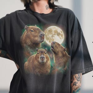 May include: Dark gray t-shirt featuring a graphic of three capybaras with a full moon in the background. The capybaras are depicted in shades of brown, with one howling at the moon. The background has a galaxy-like design.