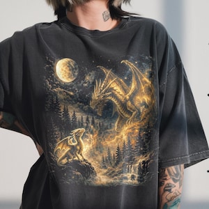 May include: Dark gray t-shirt featuring a fantasy scene with two golden dragons, a full moon, and a forest. The dragons are detailed with glowing accents, set against a night sky. The shirt has a vintage, worn look.