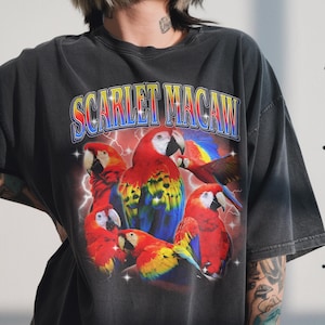 May include: Dark gray t-shirt featuring a vibrant graphic of scarlet macaws. The design includes the text "SCARLET MACAW" in a colorful, retro font, with lightning bolts and stars. The macaws are depicted in red, yellow, blue, and white.